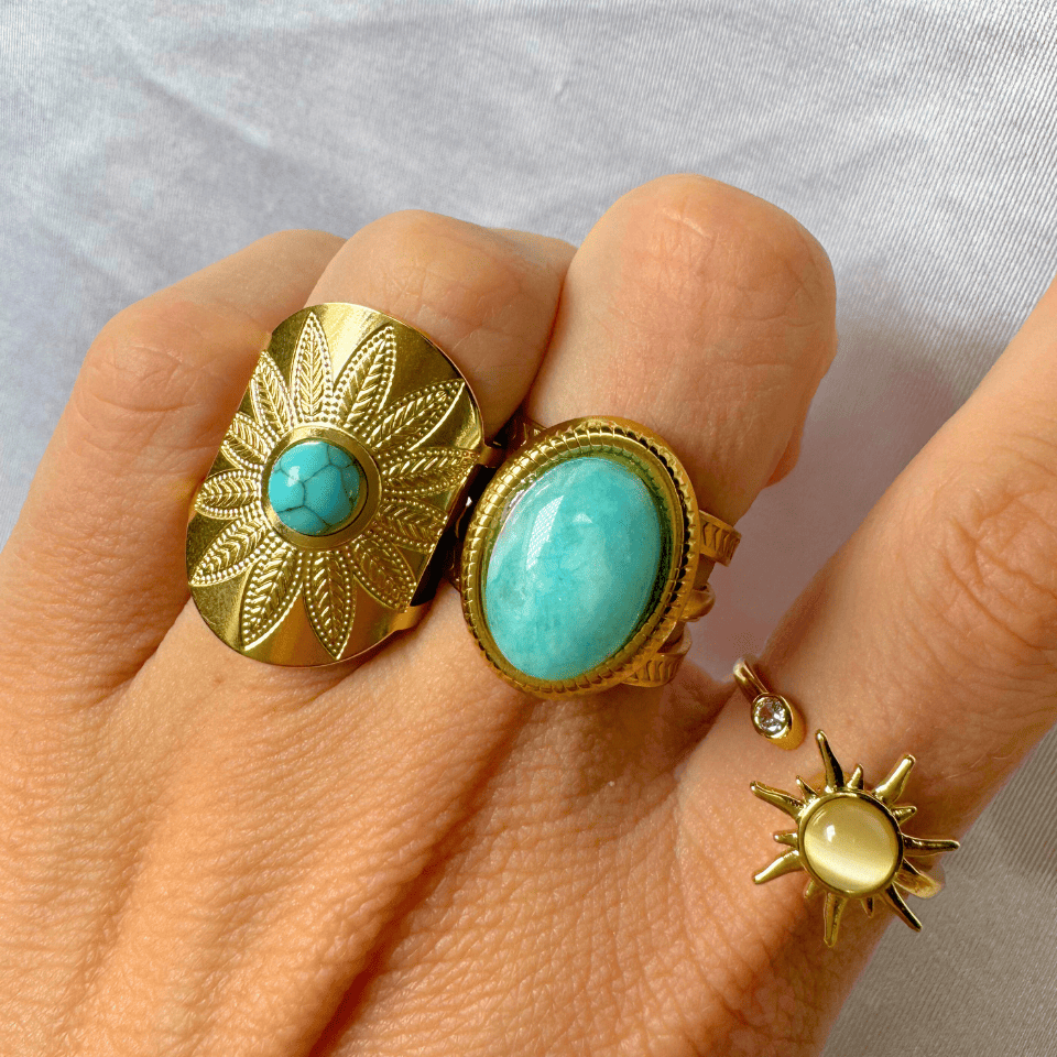 Hand wearing two gold rings with turquoise stones on a light gray background
