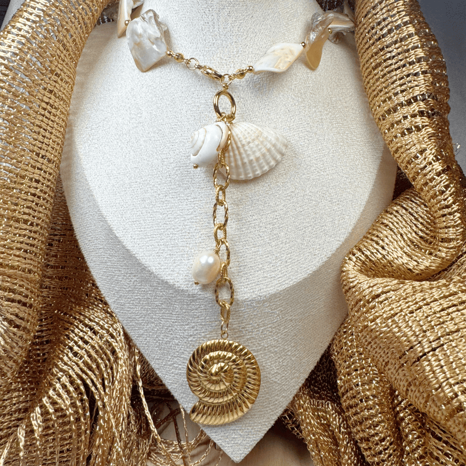 Gold necklace with shell and coin pendants on a white stand, draped with gold fabric.