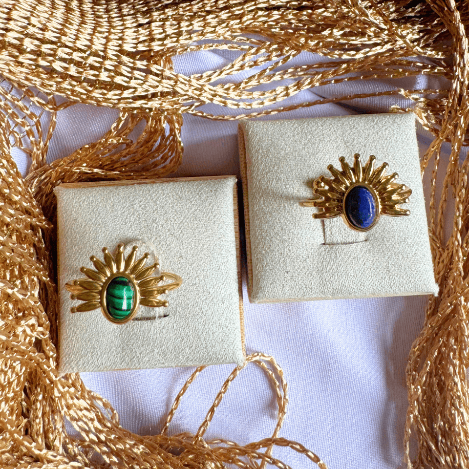 Two gold rings with gemstones on beige display boxes, surrounded by gold ribbons.