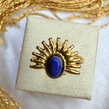 Gold ring with a blue stone on a white cushion, surrounded by gold thread.