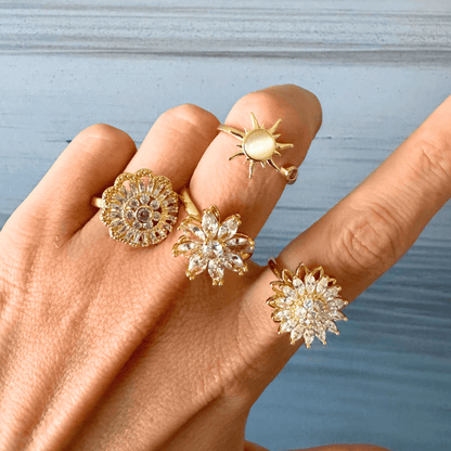 Hand wearing multiple gold rings with floral and sun designs against a light blue background