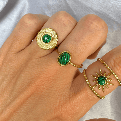 Hand wearing gold rings with green gemstones on a neutral background