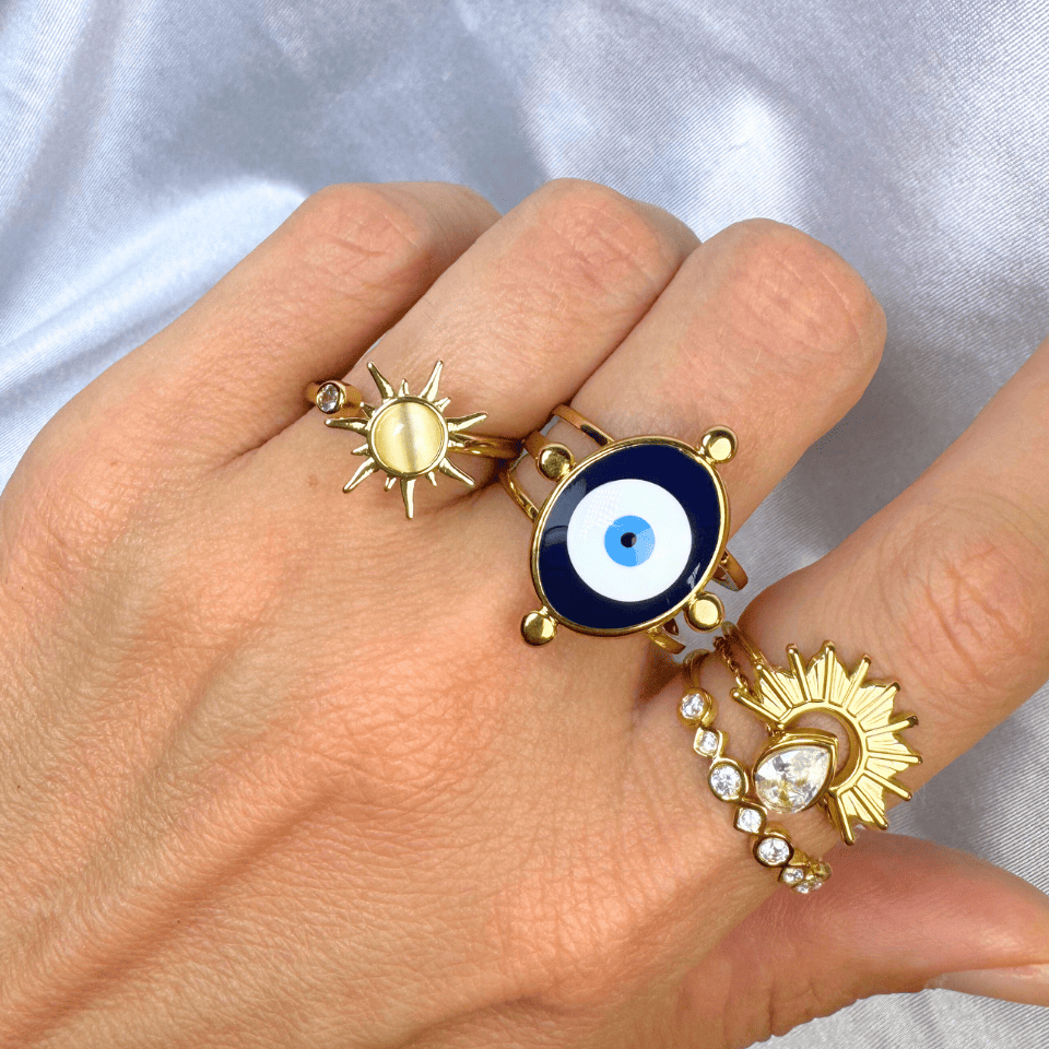 Hand wearing three gold rings with sun and eye designs on a light gray background