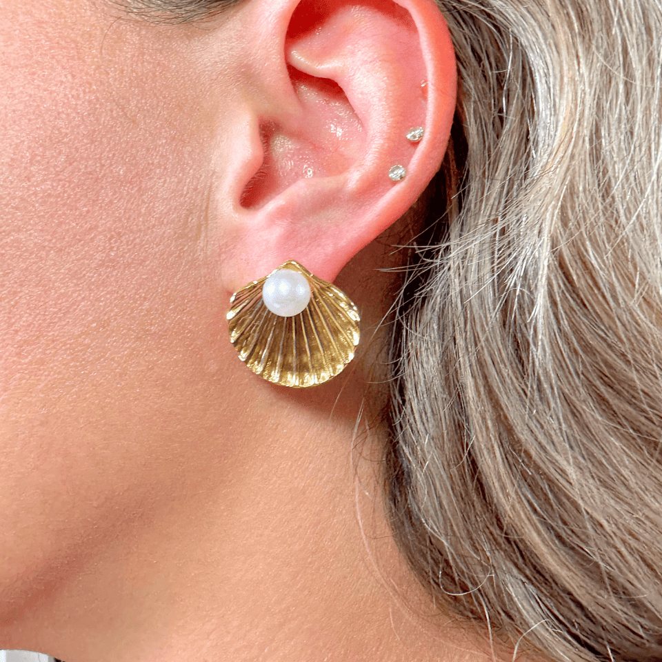 Gold shell earrings with a pearl center worn by a person with gray hair.