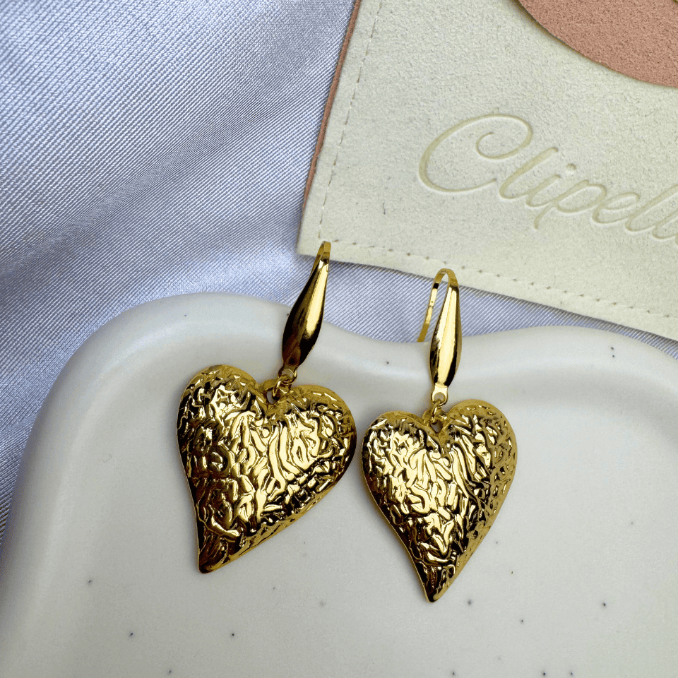 Gold heart-shaped earrings on a white dish with a brand name tag in the background.
