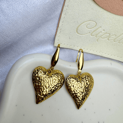 Gold heart-shaped earrings on a white dish with a brand name tag in the background.