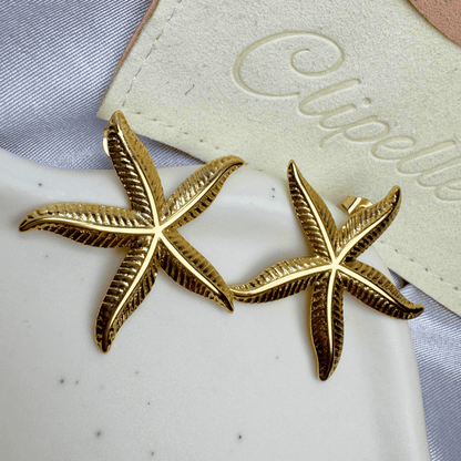 Gold starfish earrings on a white surface with a branded card in the background.
