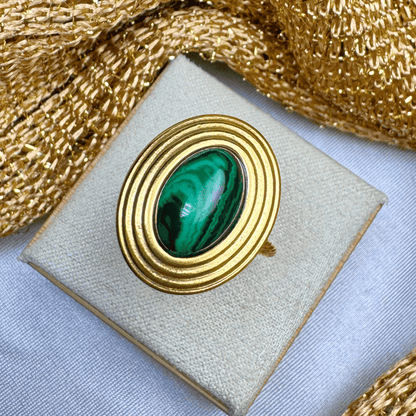 Gold brooch with green gemstone on a beige cushion, gold ribbon in the background