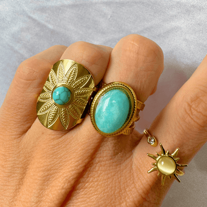 Hand wearing two gold rings with turquoise stones on a light gray background