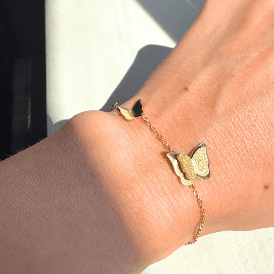 Gold bracelet with butterfly charm on a wrist against a neutral background